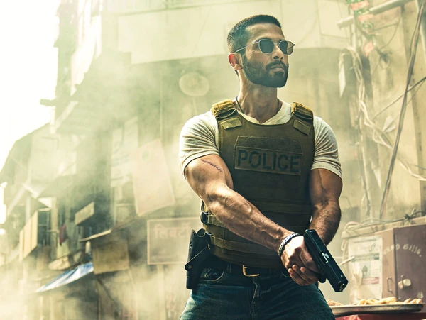 Super - cop Dev, wearing a police vest, stands alert on a dusty street in the movie "Deva".